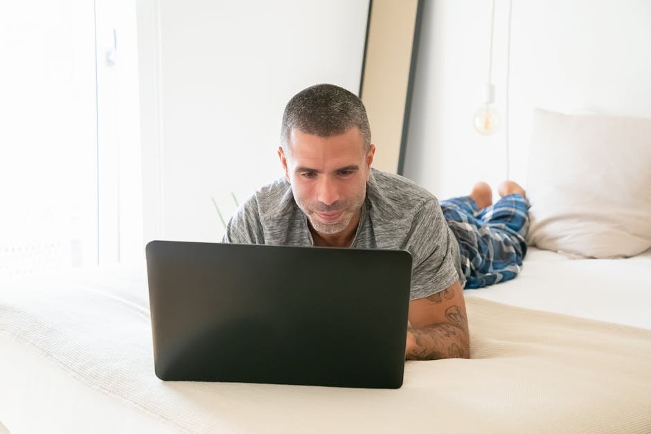 Adult man in casual attire using laptop on bed for remote work in bright home setting.