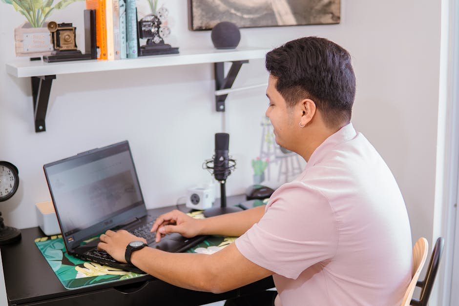 A young man sits at a desk working on his laptop, with a microphone nearby in a home office setting.