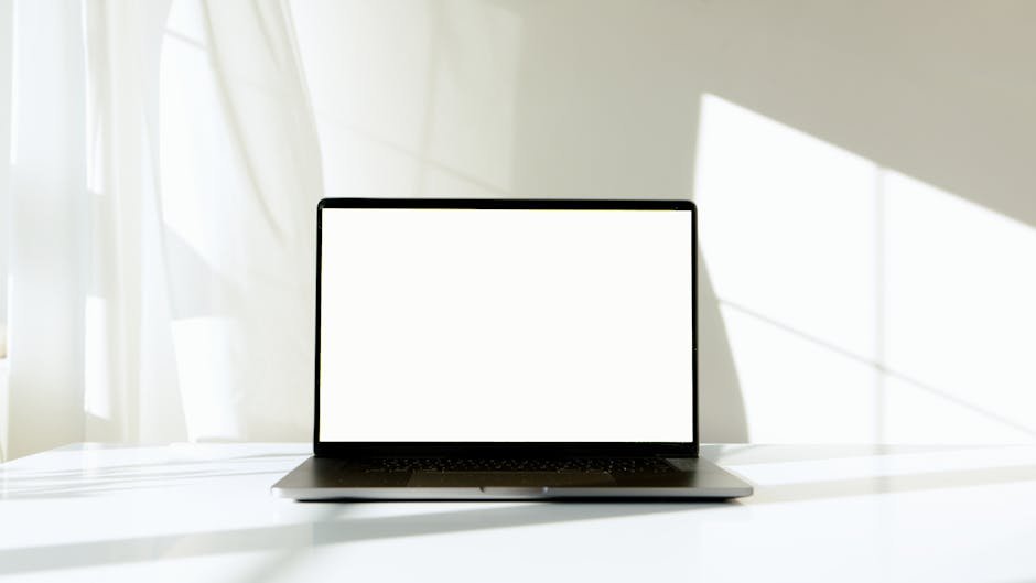 A minimalist laptop with a blank screen on a white table, bathed in natural sunlight.