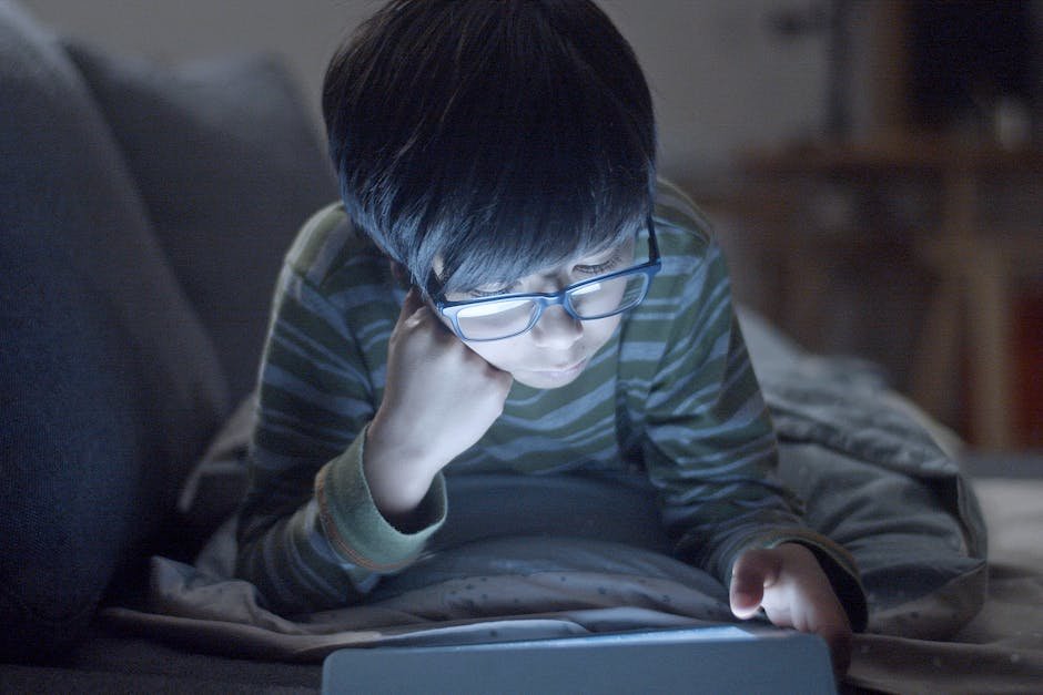 Young boy with glasses using a tablet at night, lying indoors with striped shirt.