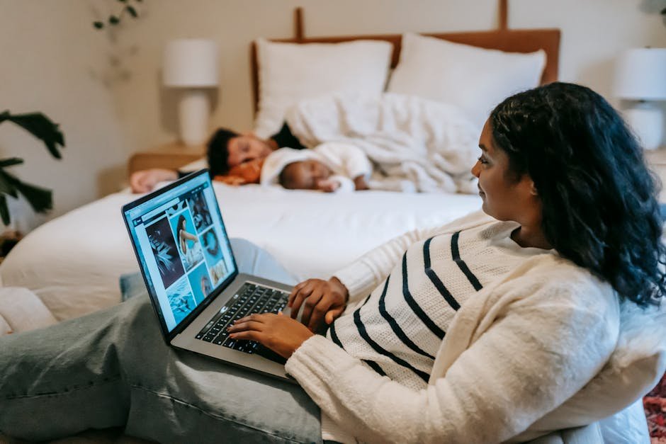 A woman uses a laptop in a cozy bedroom, with a baby sleeping on the bed nearby.