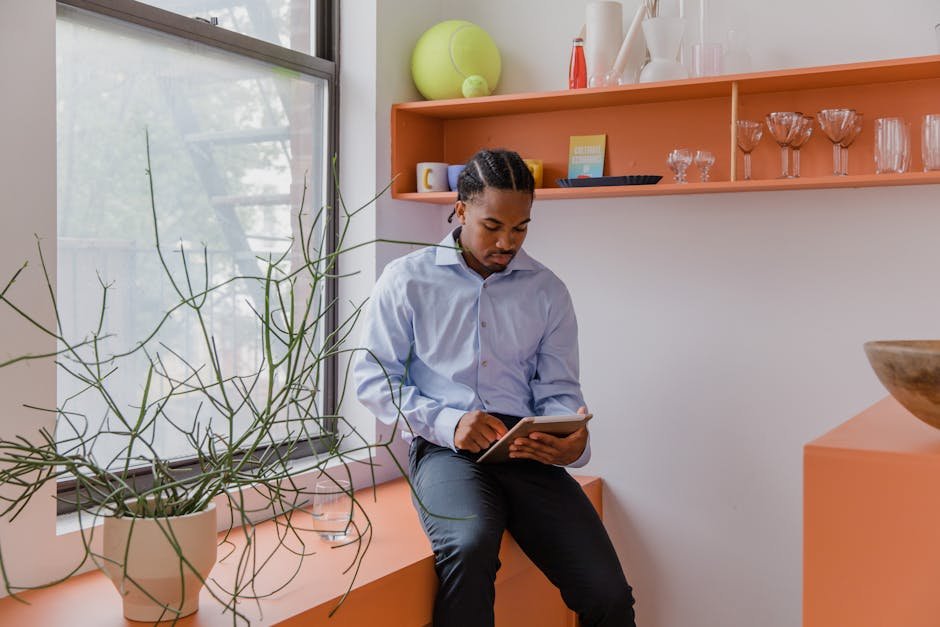 Young man sits in a modern, bright interior using a tablet, with plants and decorative items around.