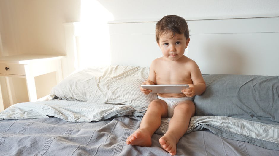 A cute baby sitting on bed with tablet, enjoying a relaxed indoor moment.